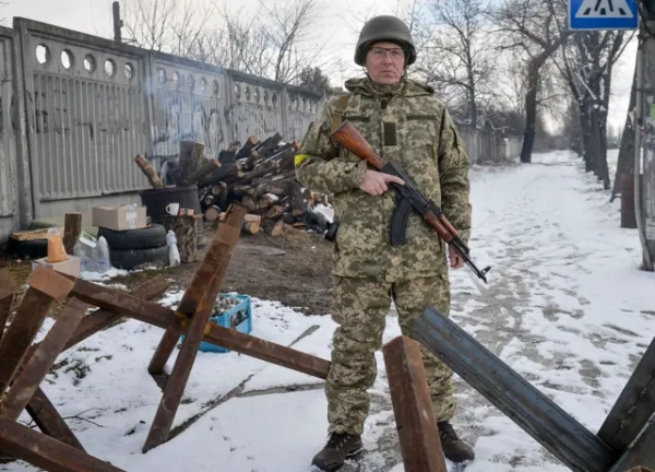 Парубій у військовій формі з жовтою стрічкою на плечі тримає в руках автомат. Він стоїть поміж протитанкових їжаків, за ним дрова і дим з багаття. Земля усипана снігом