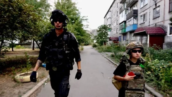 Ліворуч крокує високий чоловік у чорній формі, шоломі з навушниками та темних окулярах, із рацією та спорядженням на жилеті. Праворуч – молодша на вигляд дівчина в камуфляжному бронежилеті, шоломі та окулярах, у руках тримає червоний предмет (схоже на телефон або рацію).Вони йдуть вузькою асфальтованою дорогою між старими багатоповерхівками з балконами та зарослою зеленню обабіч
