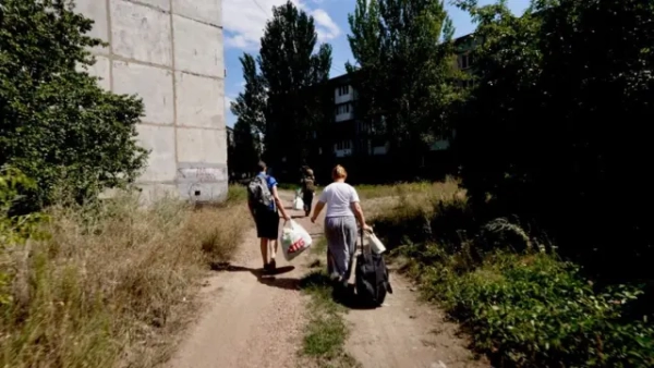 Вузькою стежкою між багатоповерховими будинками та густими кущами йдуть кілька людей із сумками та пакунками в руках. Попереду видно постать у військовому бронежилеті та шоломі, за нею йдуть жінка з великою сумкою на коліщатках і чоловік із пластиковим пакетом. Ліворуч у кадрі – стіна багатоповерхівки, праворуч – зарослі трави й кущі