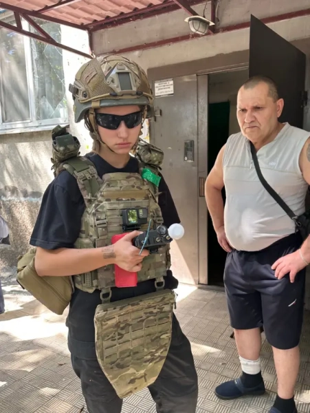 Молода дівчина у військовому спорядженні стоїть перед під’їздом багатоповерхівки. На бронежилеті закріплена камера GoPro. У руках тримає невеликий пристрій з антеною. Поруч із нею стоїть літній чоловік у спортивному одязі – світлій майці та чорних шортах, з перев’язаною гомілкою й сумкою через плече. Вони перебувають біля відкритих дверей під’їзду, на тлі видно бетонну стіну з вікном і поламаним склом.