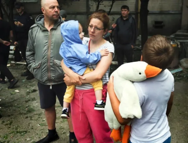 Жінка в рожевих штанях тримає дитину в синій куртці. Хлопчик 9-10 років тримає м'яку іграшку гусака та стоїть спиною до камери. Чоловік у зеленій куртці дивиться вбік.