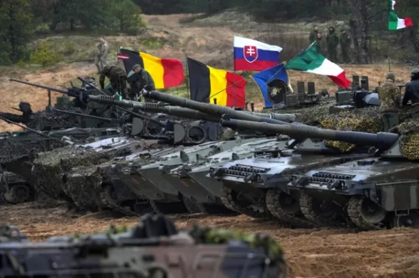 Ряд сучасних бойових танків стоїть на полі під час військових навчань. На баштах прикріплені національні прапори Бельгії, Словаччини, Італії та інших країн НАТО. Солдати в камуфляжі працюють біля техніки, на задньому плані серед дерев солдати.