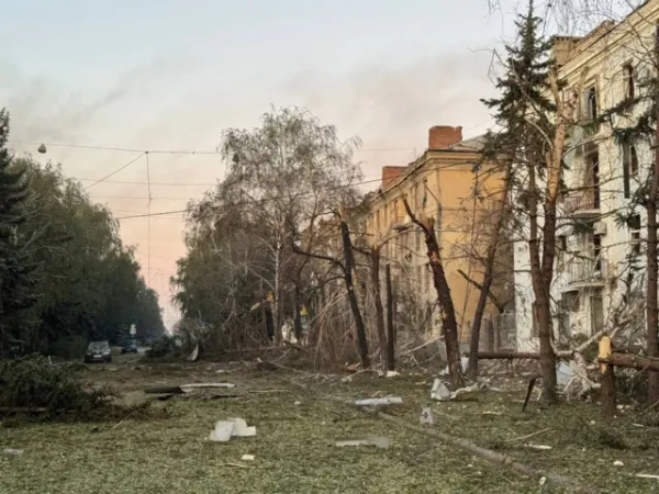 Вулиця повністю вкрита листям та гілками дерев, зрубаними осколками авіабомб. Від кількох дерев вздовж дороги залишилися лише стовбури та пні, кілька впали на землю. Усі вікна на трьох поверхах багатоквартирного будинку розбиті, рами перекошені. На задньому плані припарковані автомобілі.