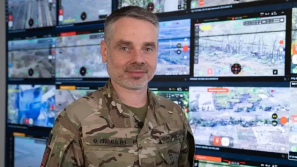 Юрій Мироненко стоїть у військовій формі перед стіною екранів, на яких зображено поля, кущі та кілька будинків. Усі сцени з російсько-українського фронту.
